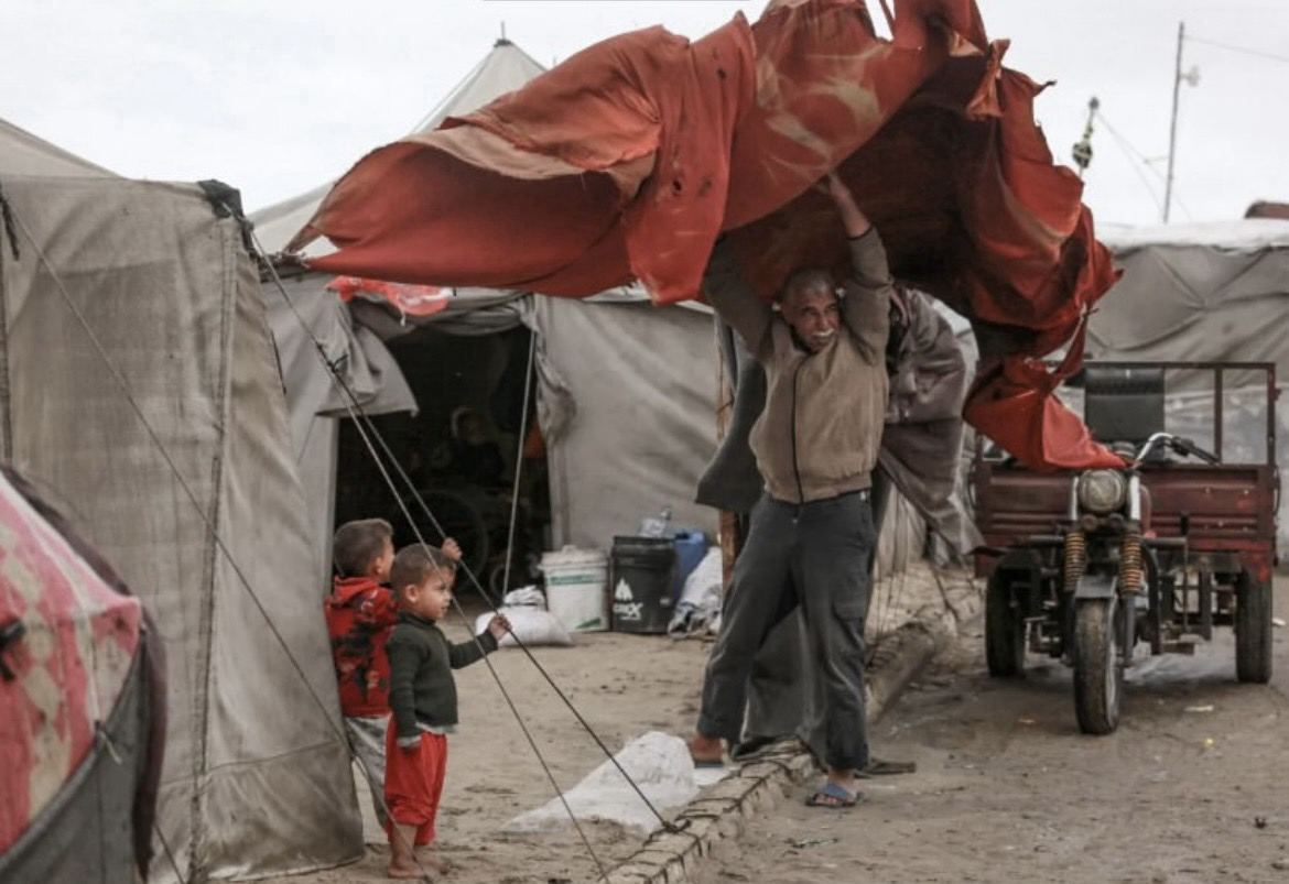 A older Palestinian man holds a tent, securing it from the wind, while two young children watch